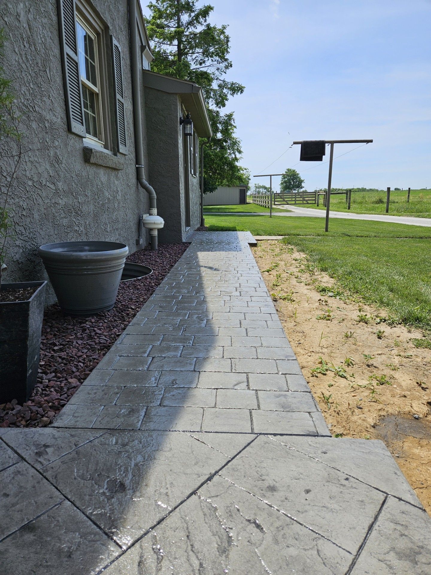 Stone walkway beside a house, bordered by landscaping, leading toward a grassy roadside field.