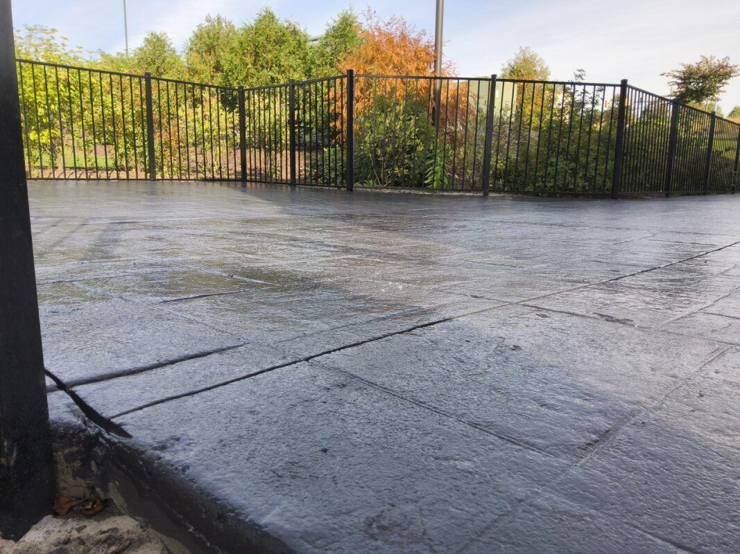 Dark, wet patio with a black metal fence and green trees in the background.