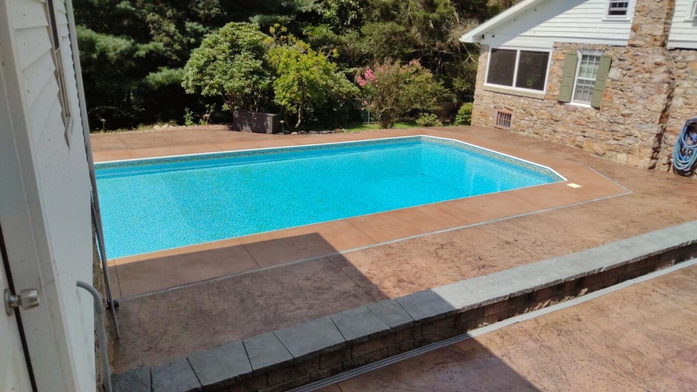 Poolside view of a rectangular pool with turquoise water, brown concrete patio, and a stone house in the background.