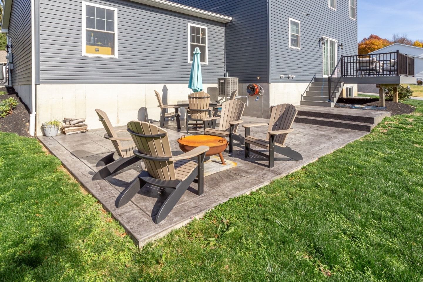 Backyard patio with seating around a fire pit, next to a gray house with green grass.