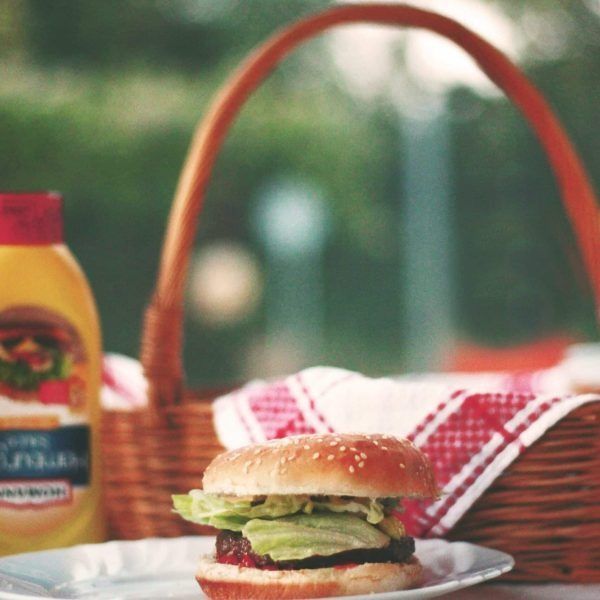 Hamburger on a plate, picnic basket with red and white cloth, mustard bottle outdoors.