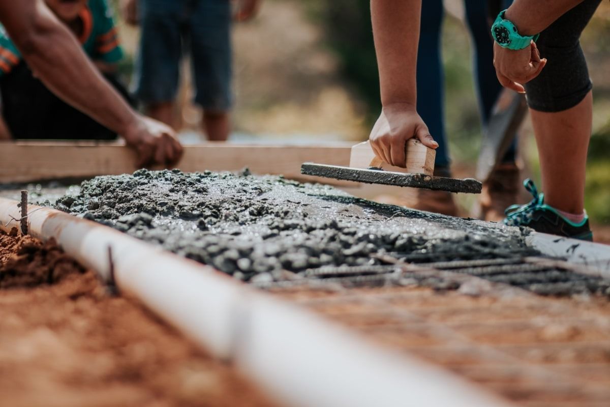Hands smoothing wet concrete with a trowel in a wooden frame; outdoor construction site.