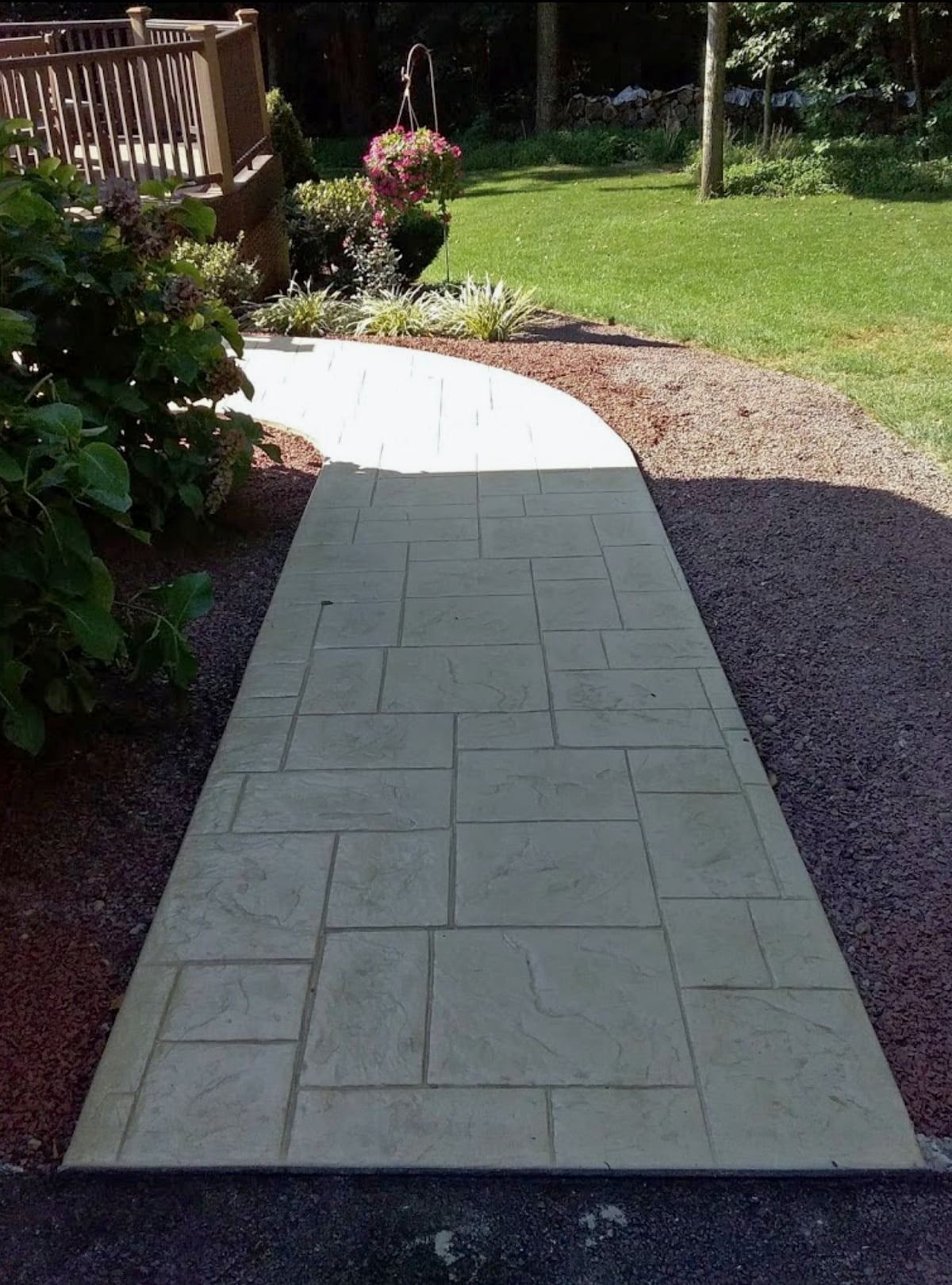 Concrete walkway with a stone pattern curves through a yard, surrounded by red gravel and grass.