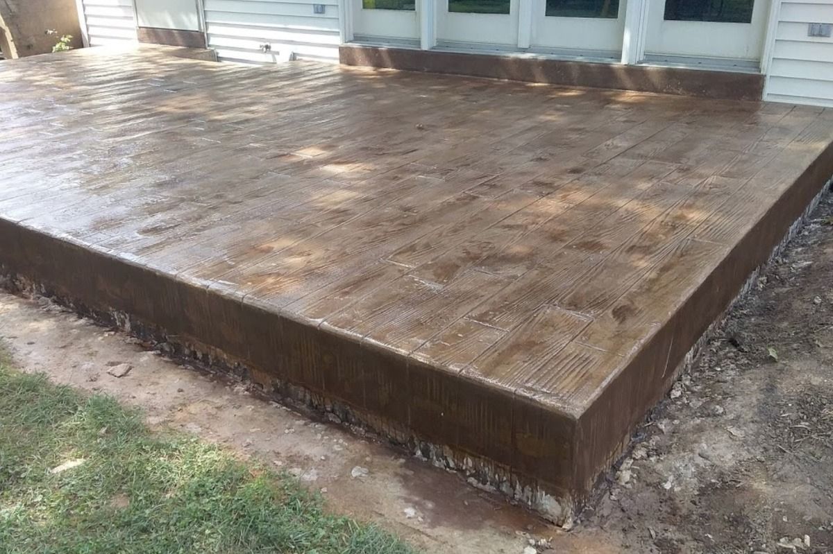 Wet, brown concrete patio next to a house, edged by dirt and grass.