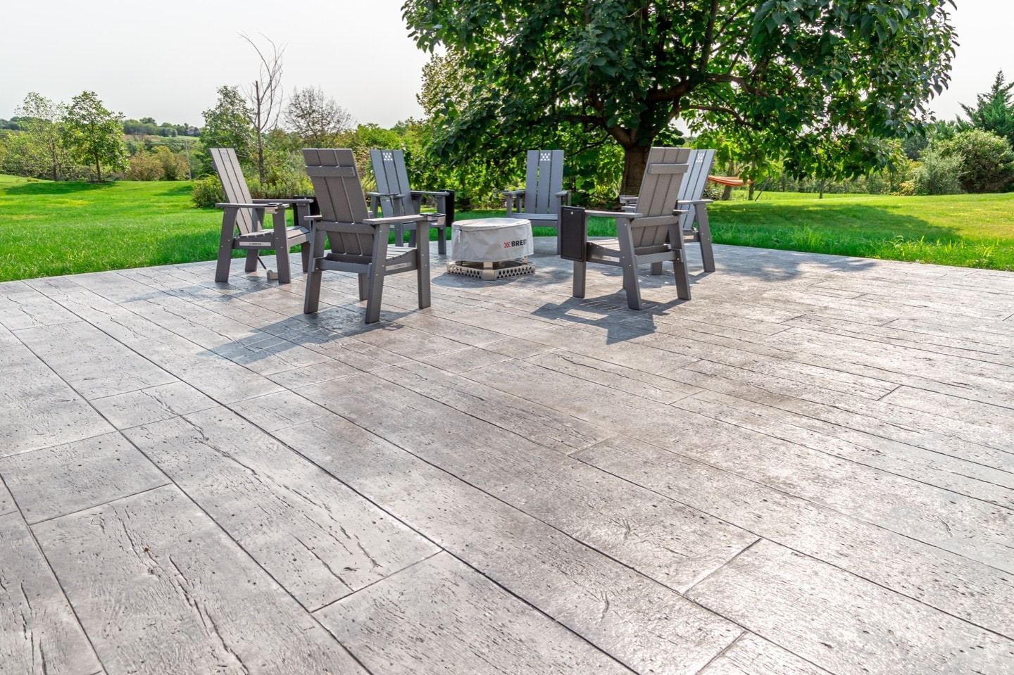 Gray patio with chairs around a fire pit, green lawn, and trees in the background on a sunny day.
