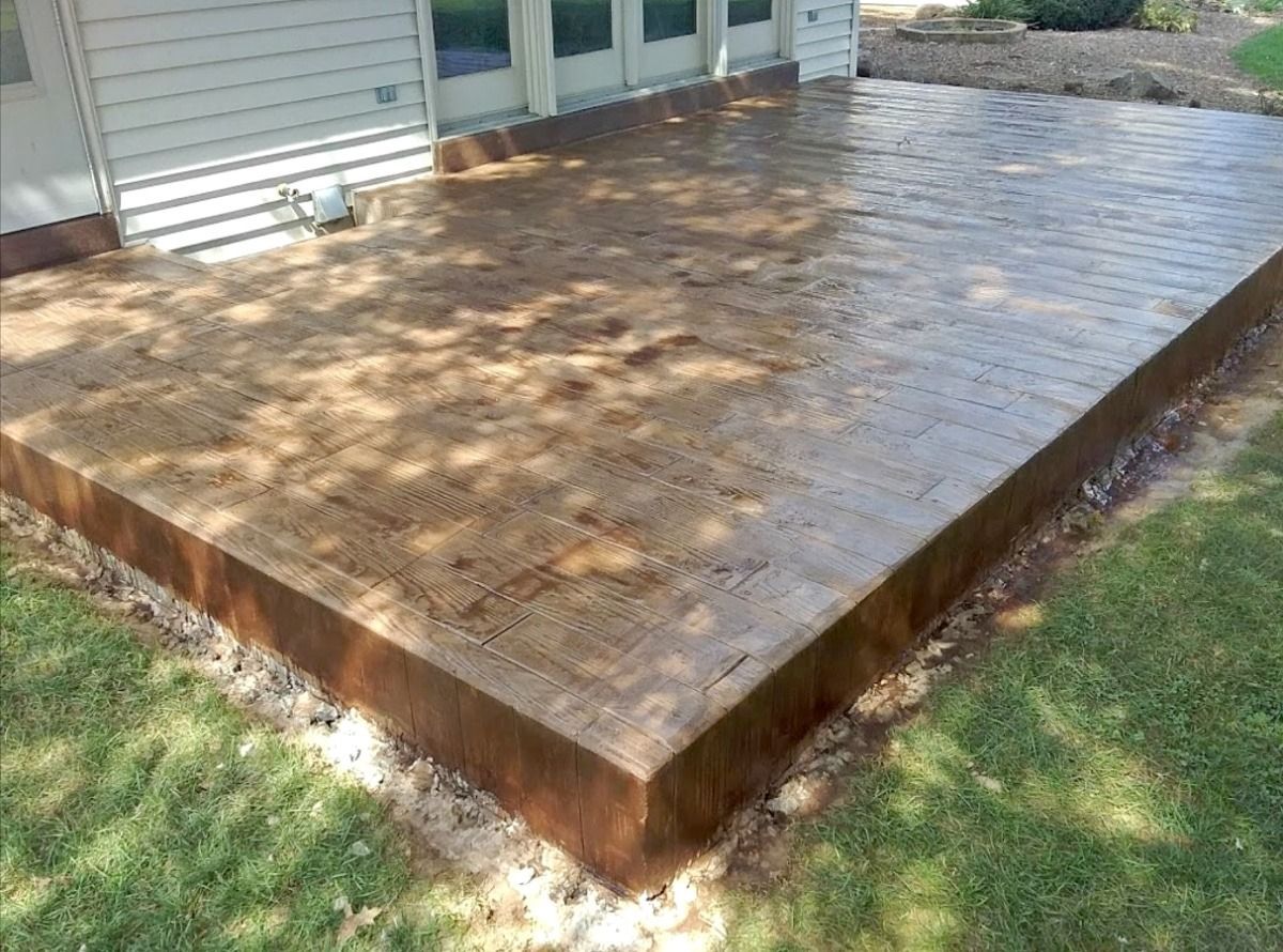 Finished concrete patio with brown edging and a house in the background. Lawn surrounds the patio.
