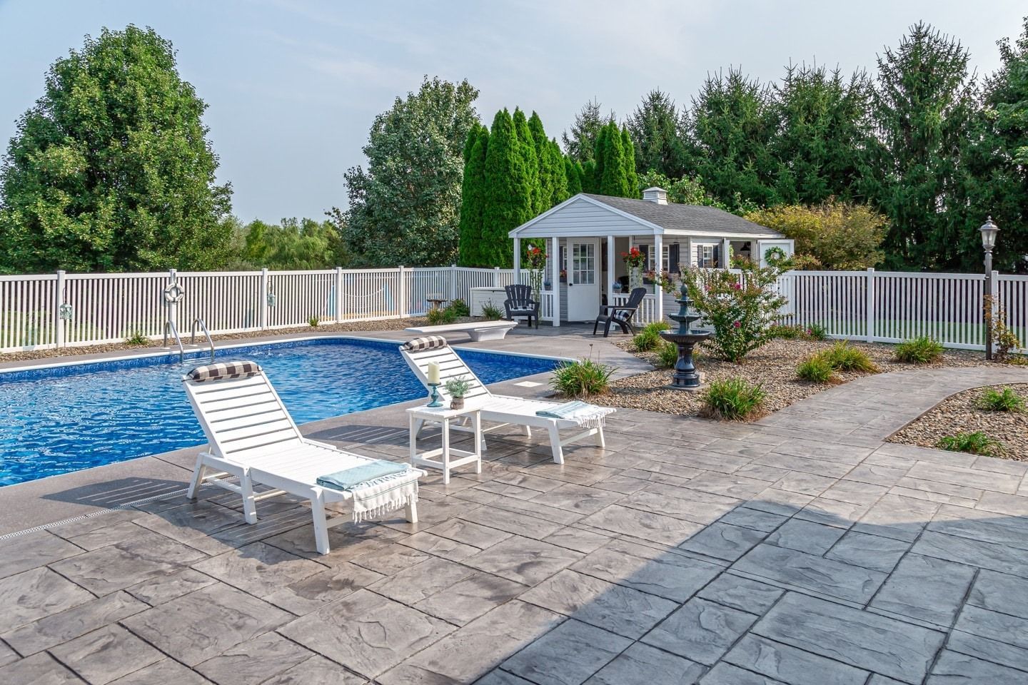 Swimming pool and lounge chairs on patio near a small white gazebo and trees on a sunny day.