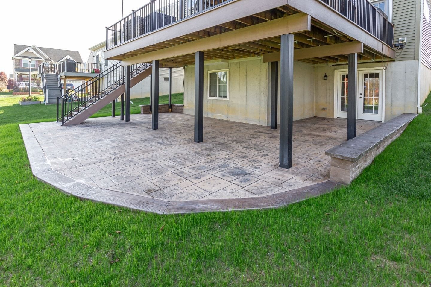 Concrete patio under a deck with green grass. A retaining wall with a bench surrounds the patio.