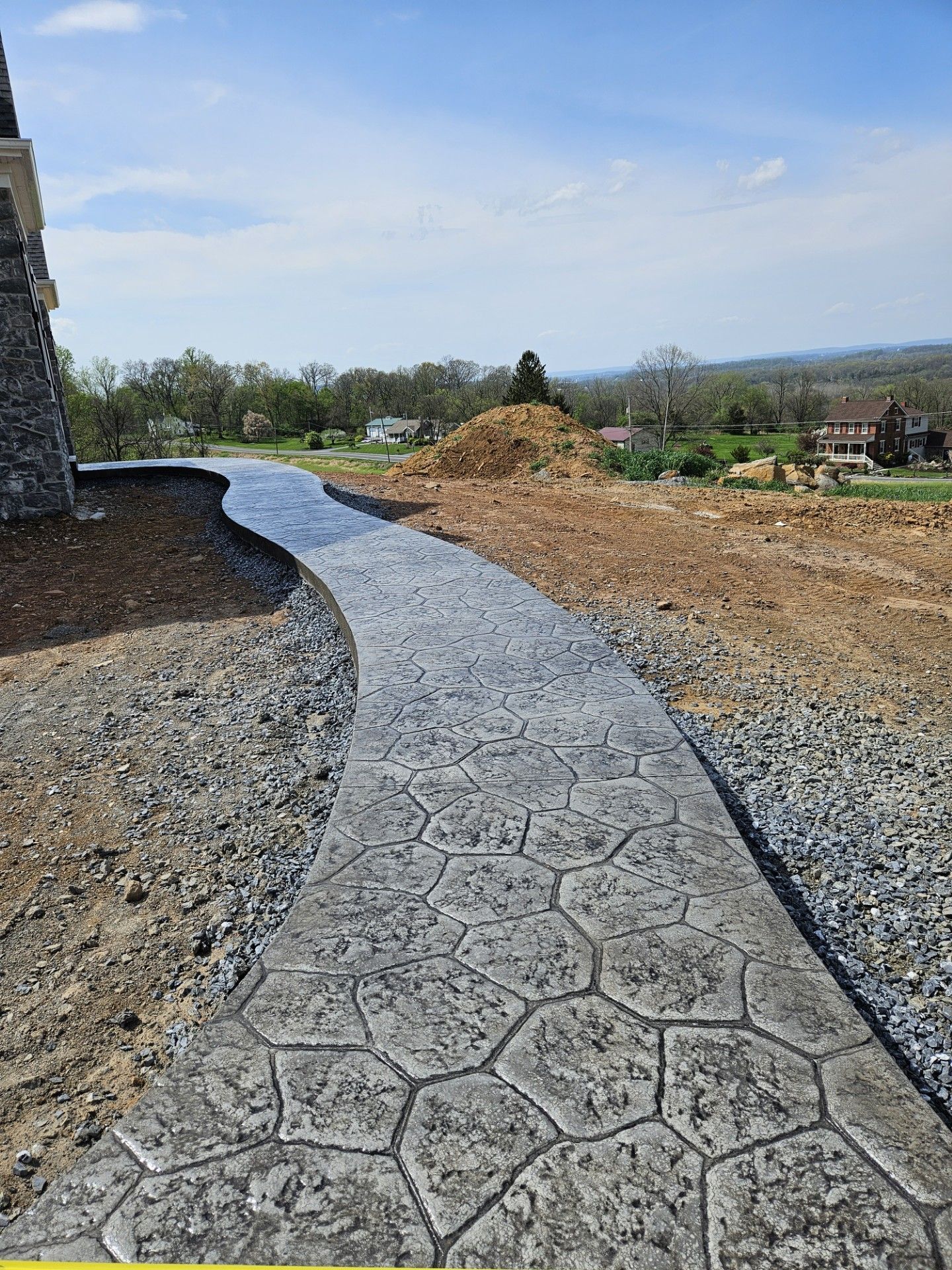 Curving stamped concrete walkway beside a stone wall, dirt pile, and open landscape under a blue sky.