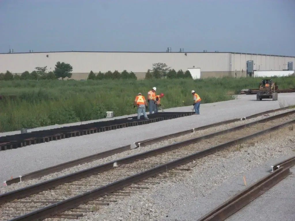 Construction workers in orange vests repairing a railroad track beside a road and grassy field.