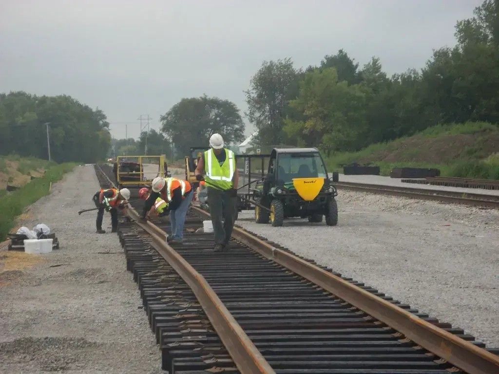 Workers repair railroad tracks beside a green rail vehicle on a cloudy day