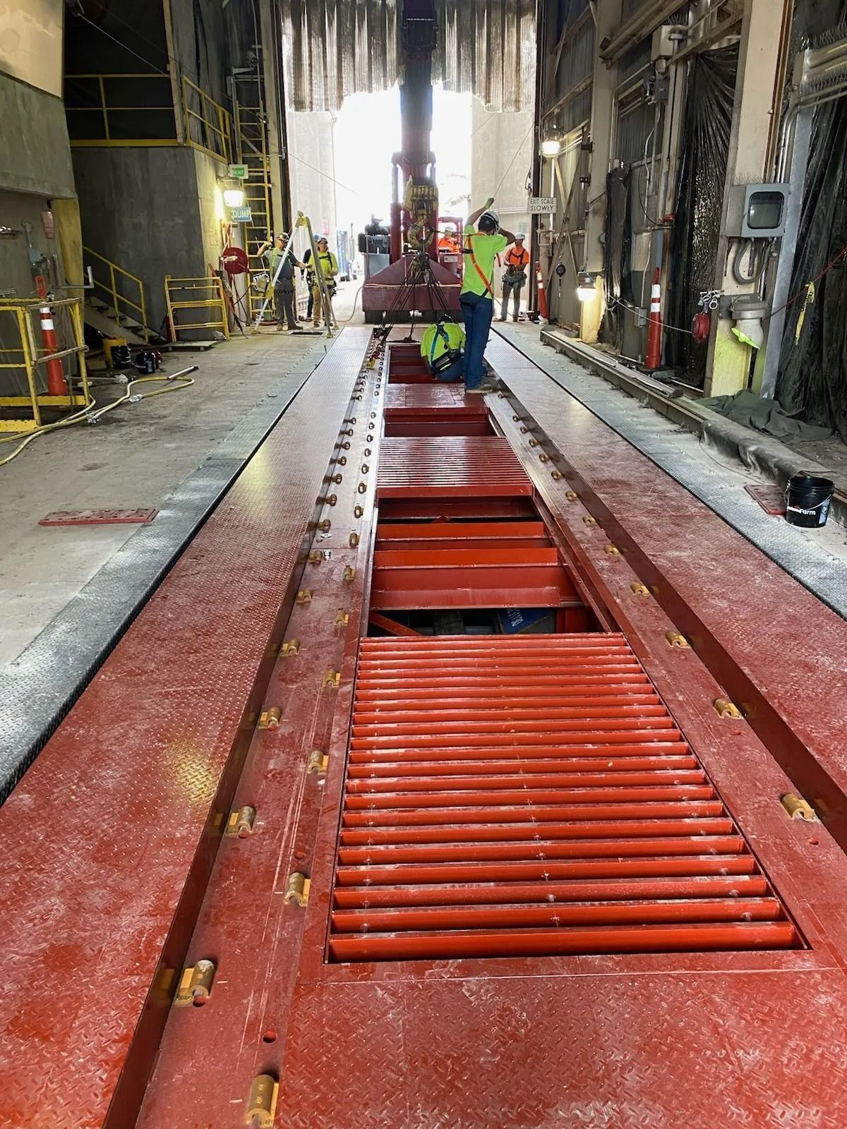 Red industrial trench with metal grates inside a worksite, with workers in safety gear in the background
