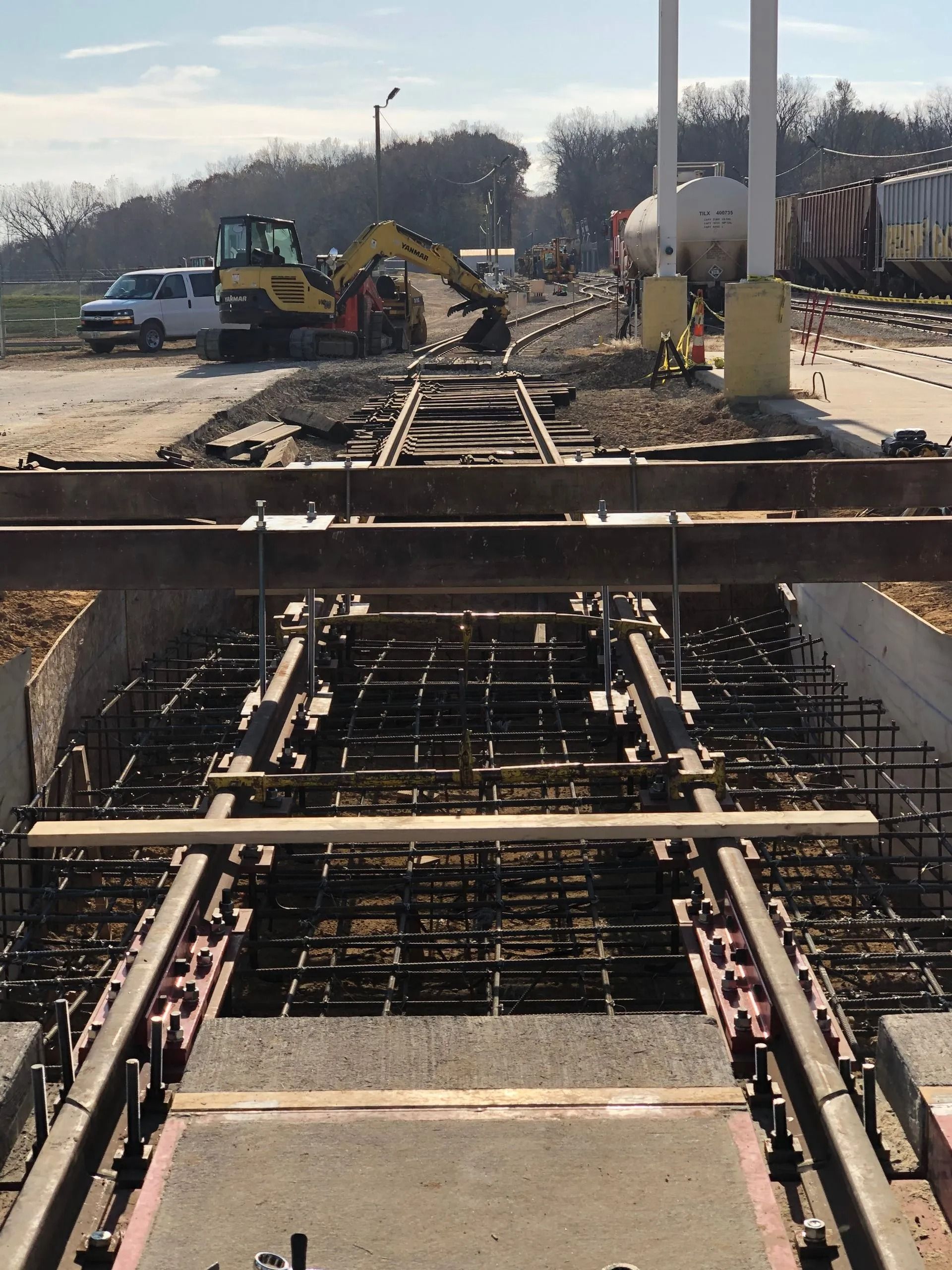 Construction site with a concrete form and rebar in foreground, excavator and trucks in the background.
