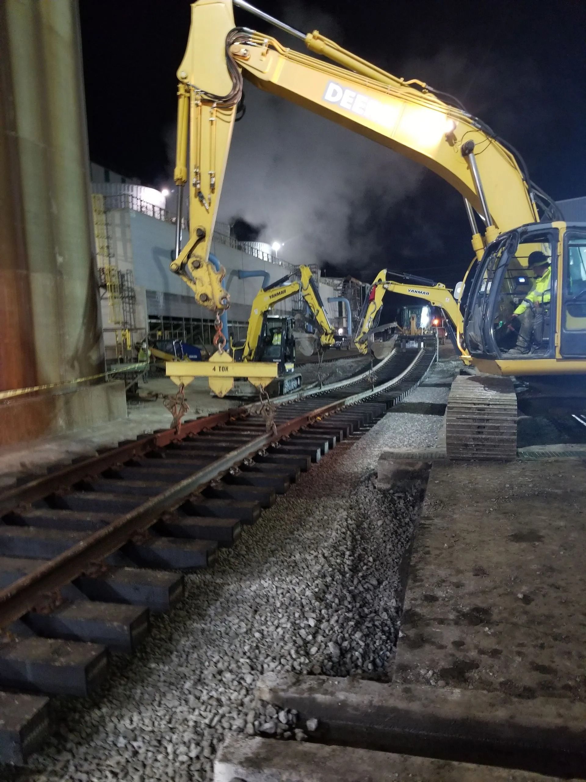 Yellow excavators working beside railroad tracks at night, with steam or smoke in the air.
