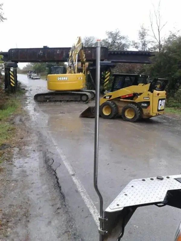 Yellow excavator and skid steer clearing debris under a low bridge on a rainy road.