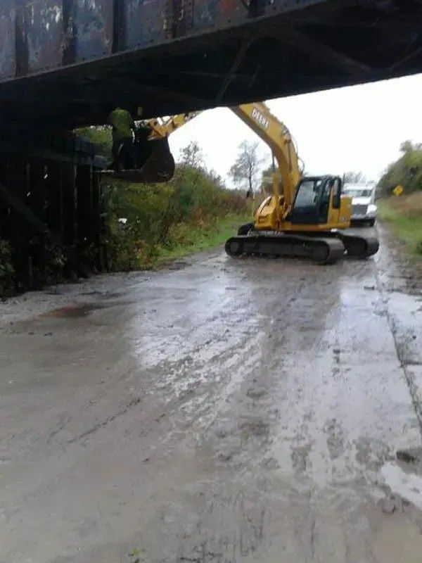 Yellow excavator clearing debris on a wet road beneath a bridge