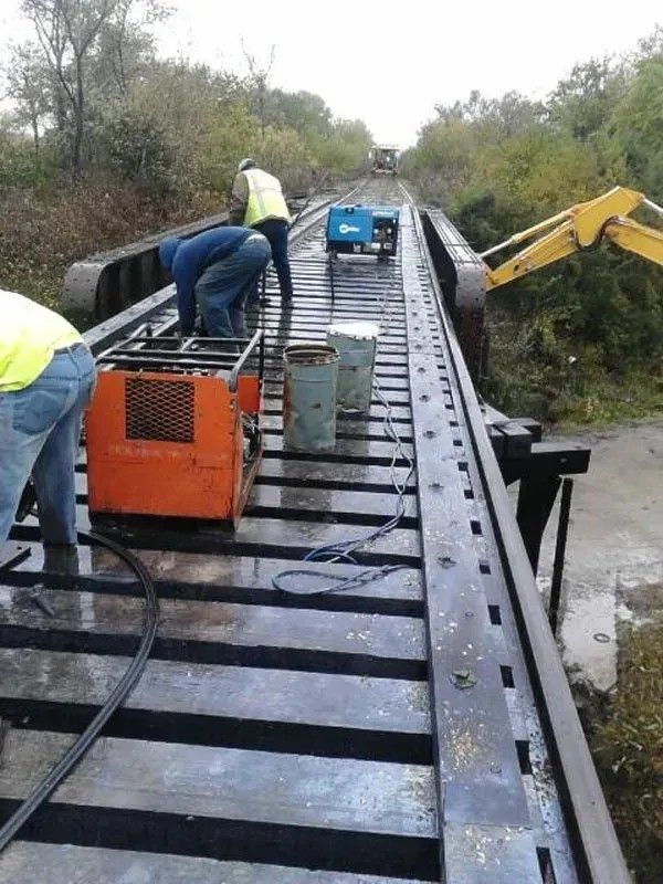 Workers repair a metal walkway with tools and machines along a raised bridge in a wooded area