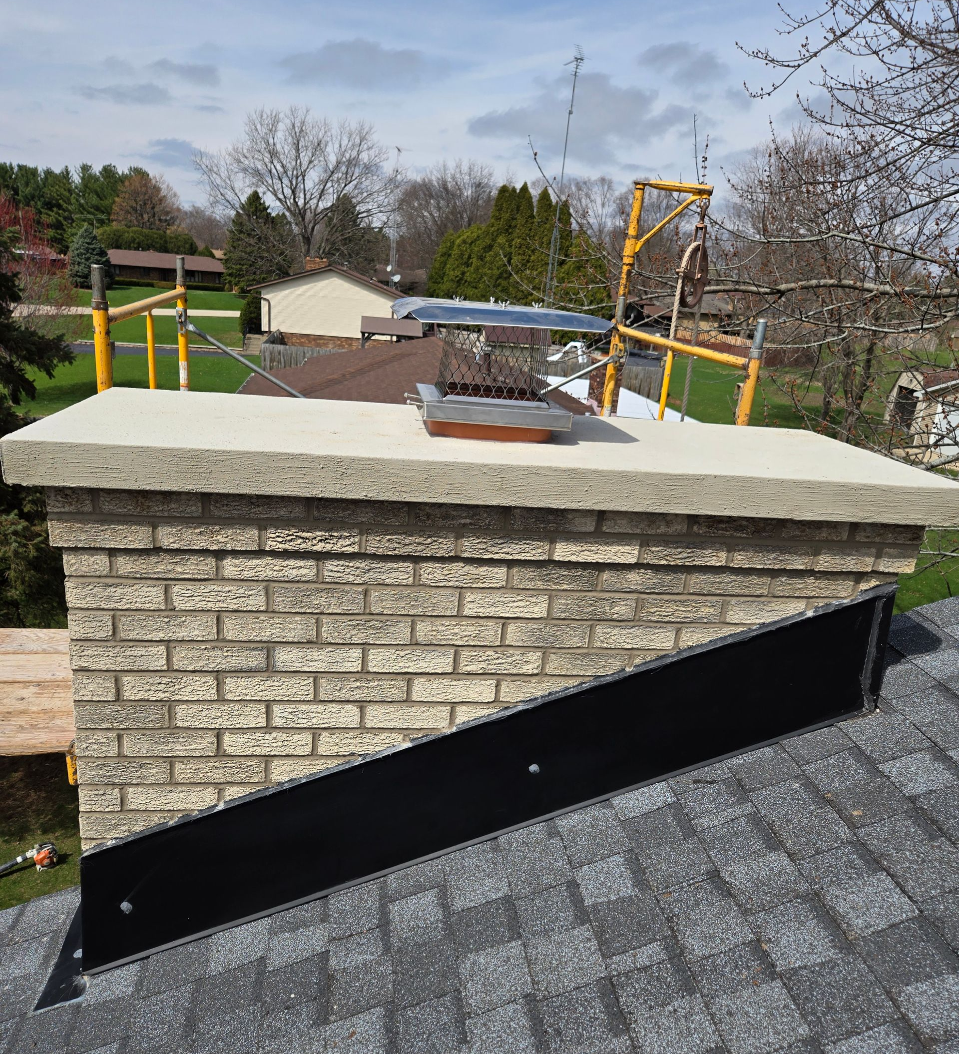 A chimney on a residential roof featuring a concrete cap, a metal flue cover, and new black flashing along the slope.