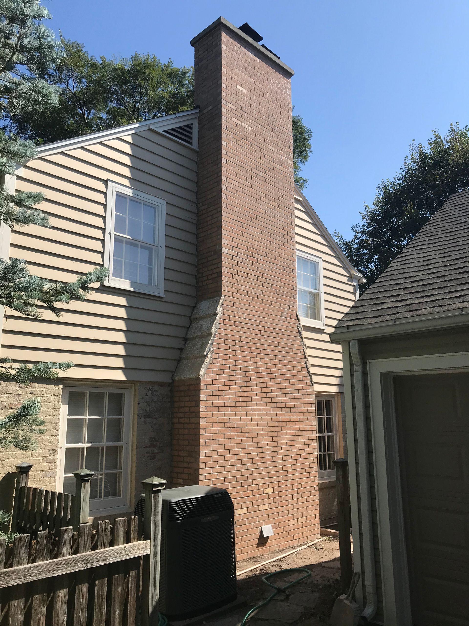 Exterior of a house featuring a tall, central brick chimney against light-colored siding, with trees under a blue sky.