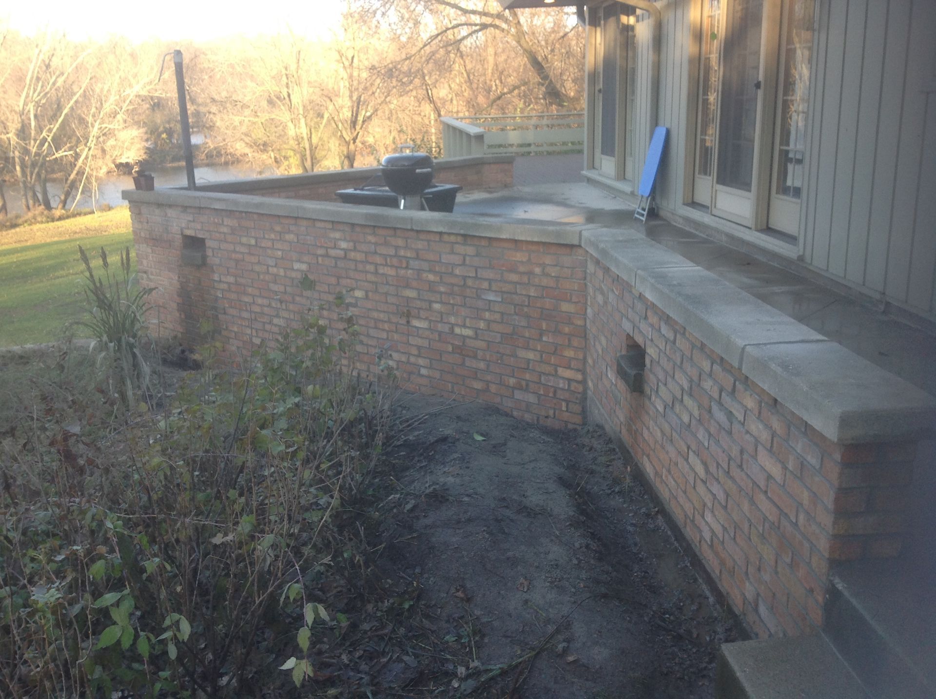 A multi-level brick retaining wall borders a patio with a grill, set against a lawn and trees in soft, golden sunlight.