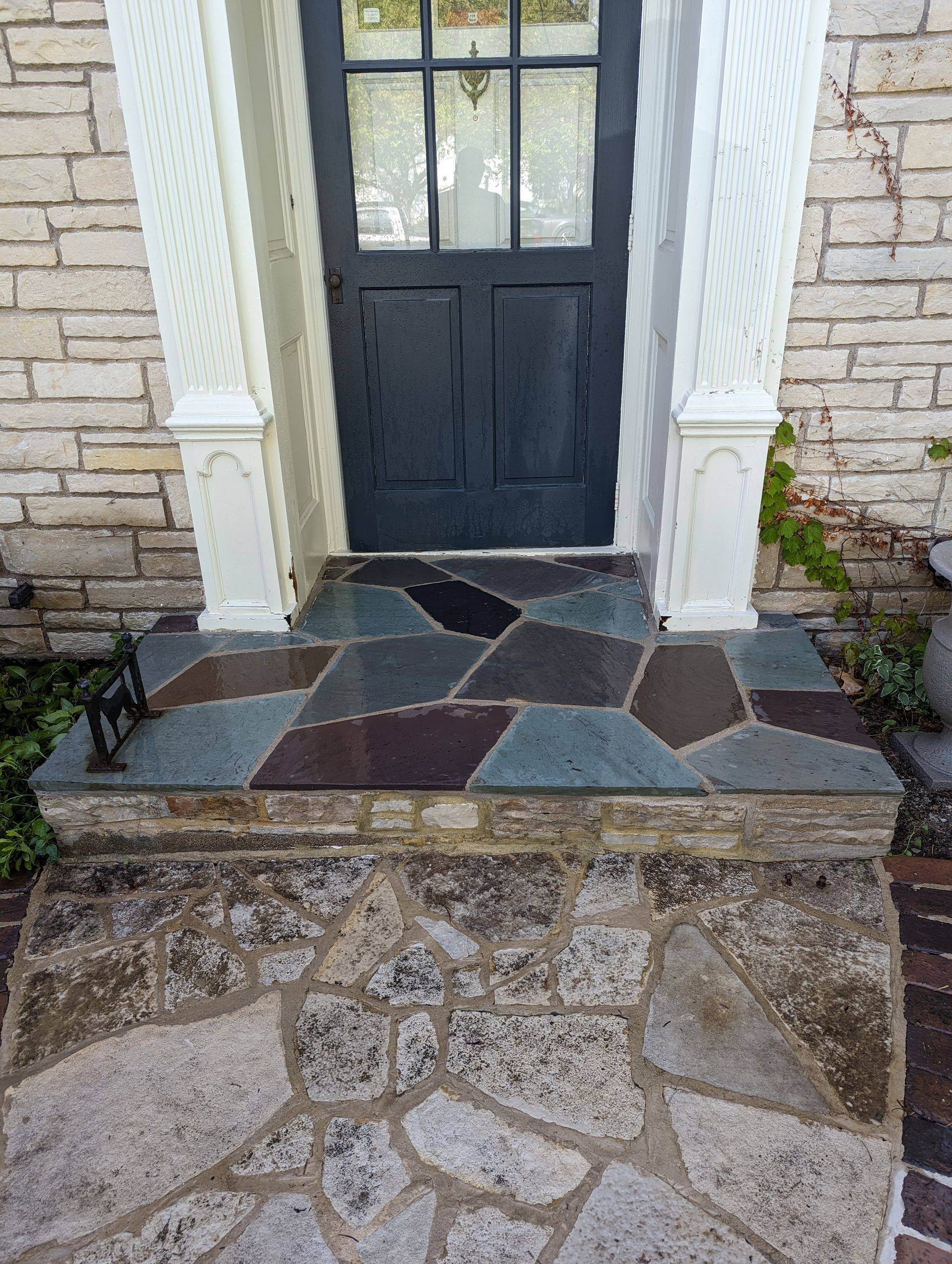 A front entrance with a dark door framed by white columns, featuring a flagstone step and a textured stone walkway.