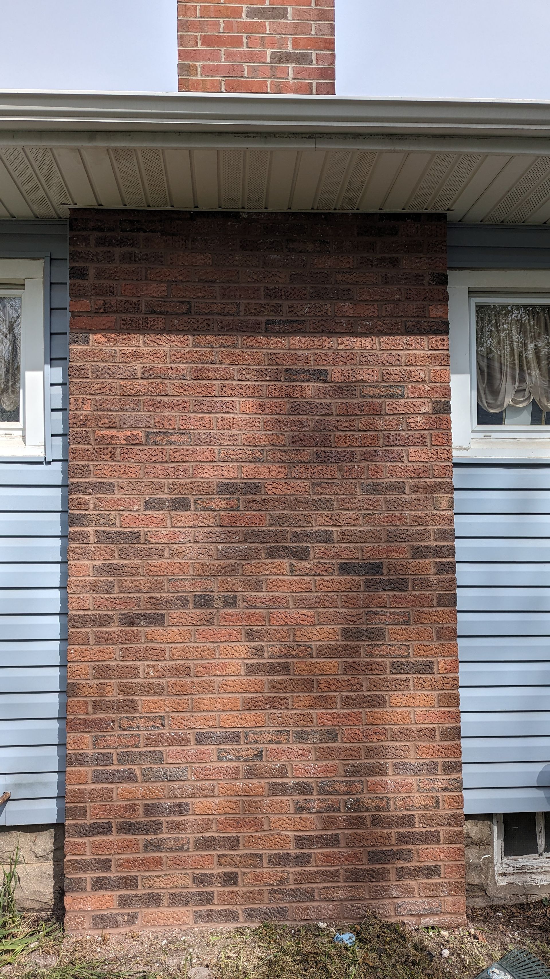 Exterior view of a rectangular brick chimney centered between two windows on a house with light blue siding.