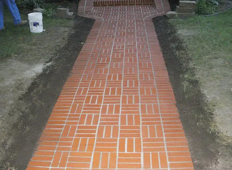 Narrow brick walkway with orange patterned pavers leading through a grassy yard.