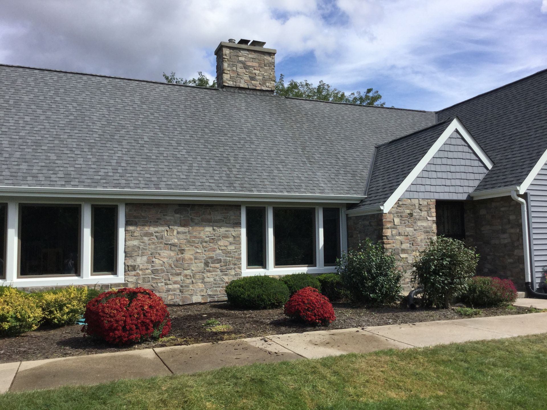 A ranch-style house with stone veneer, grey shingled roof, a stone chimney, and landscaping with red and green bushes.