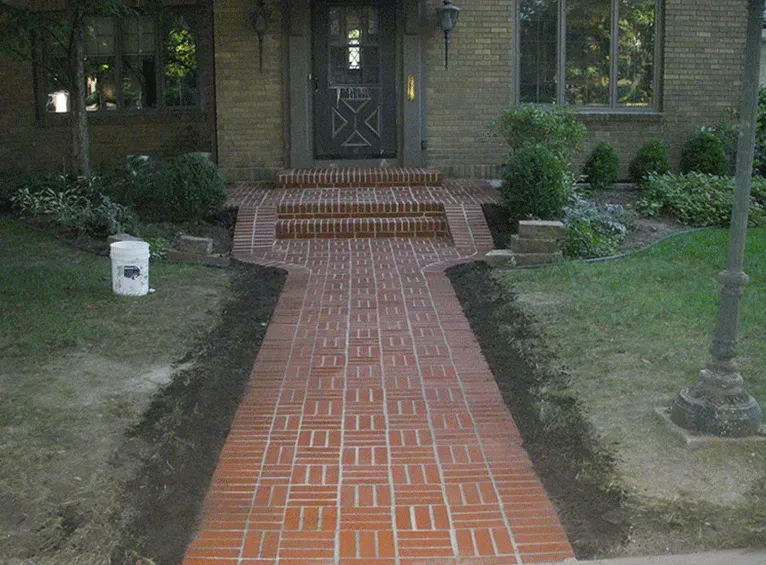 A straight red brick walkway leads to the front porch and steps of a brick house with green shrubs on either side.