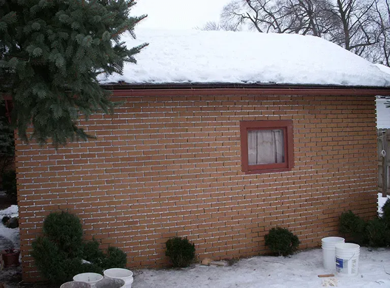 A brick detached garage with a snow-covered roof and a single window, surrounded by small bushes and a few white buckets.