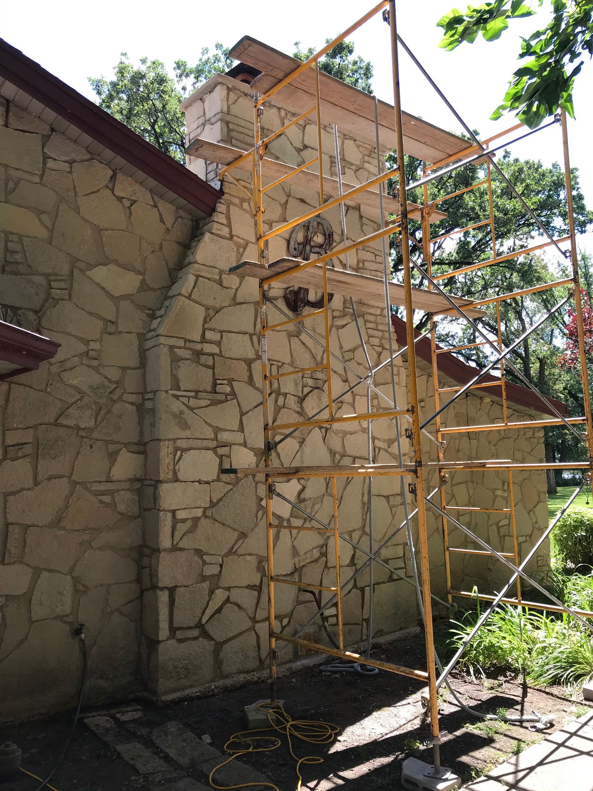A stone house chimney undergoing repair with yellow metal scaffolding surrounding it on a sunny day.