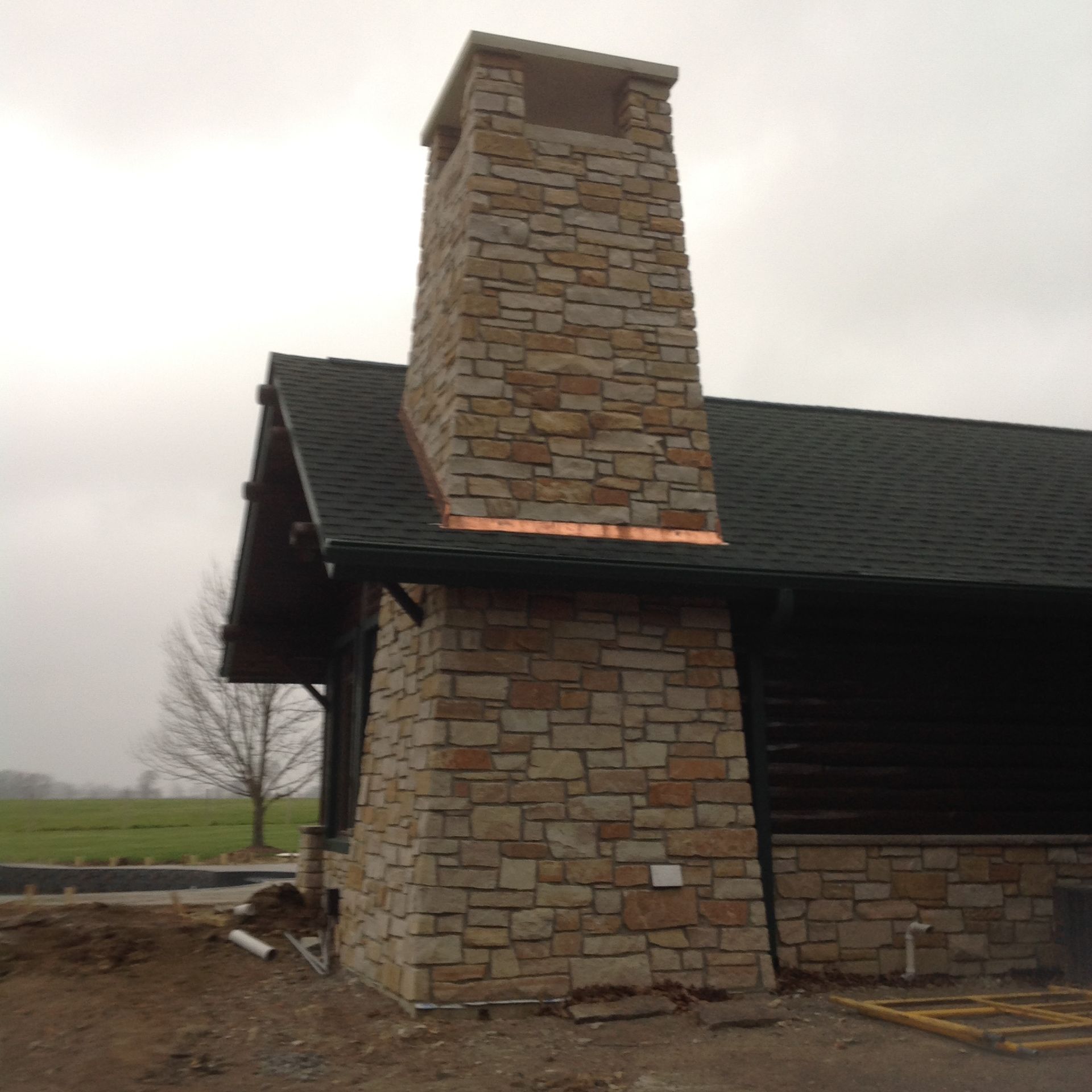 A stone chimney attached to a dark-sided building with a shingled roof, set against an overcast sky.