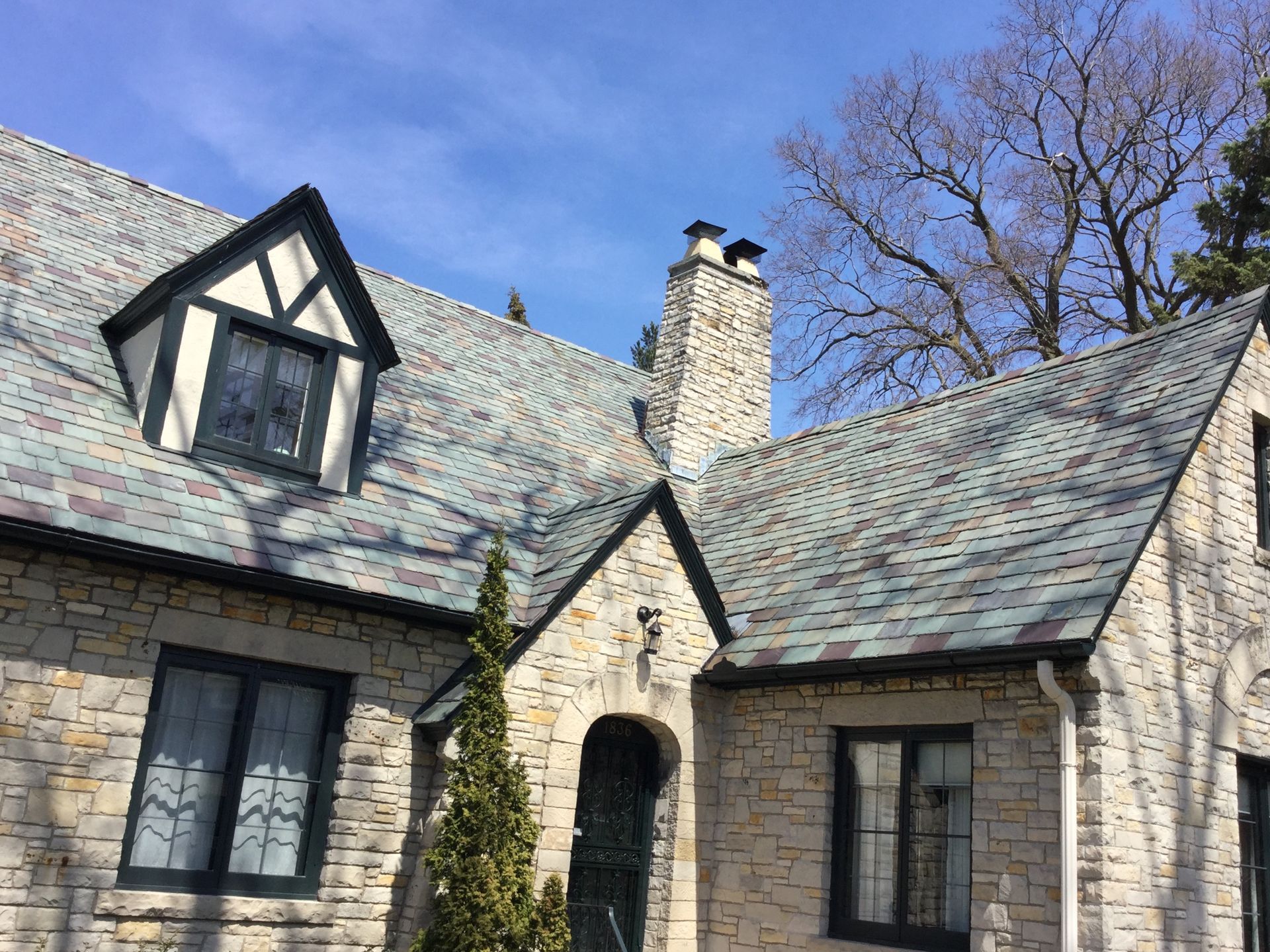 A stone cottage with dark-framed windows, a multi-textured roof, and a chimney set against a bright blue sky.