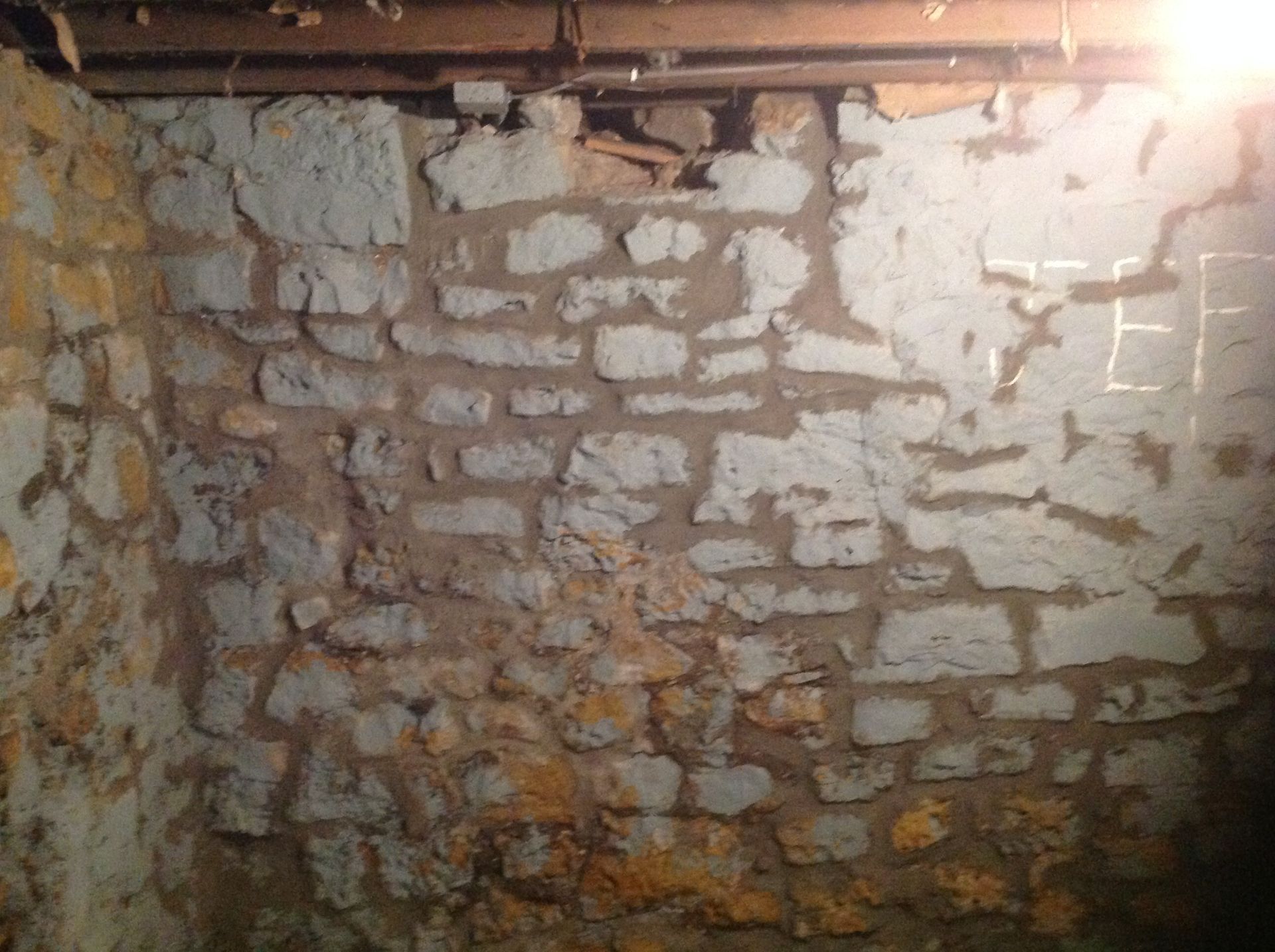 A dimly lit basement corner with rough, gray-and-tan stone walls and exposed wooden ceiling joists.