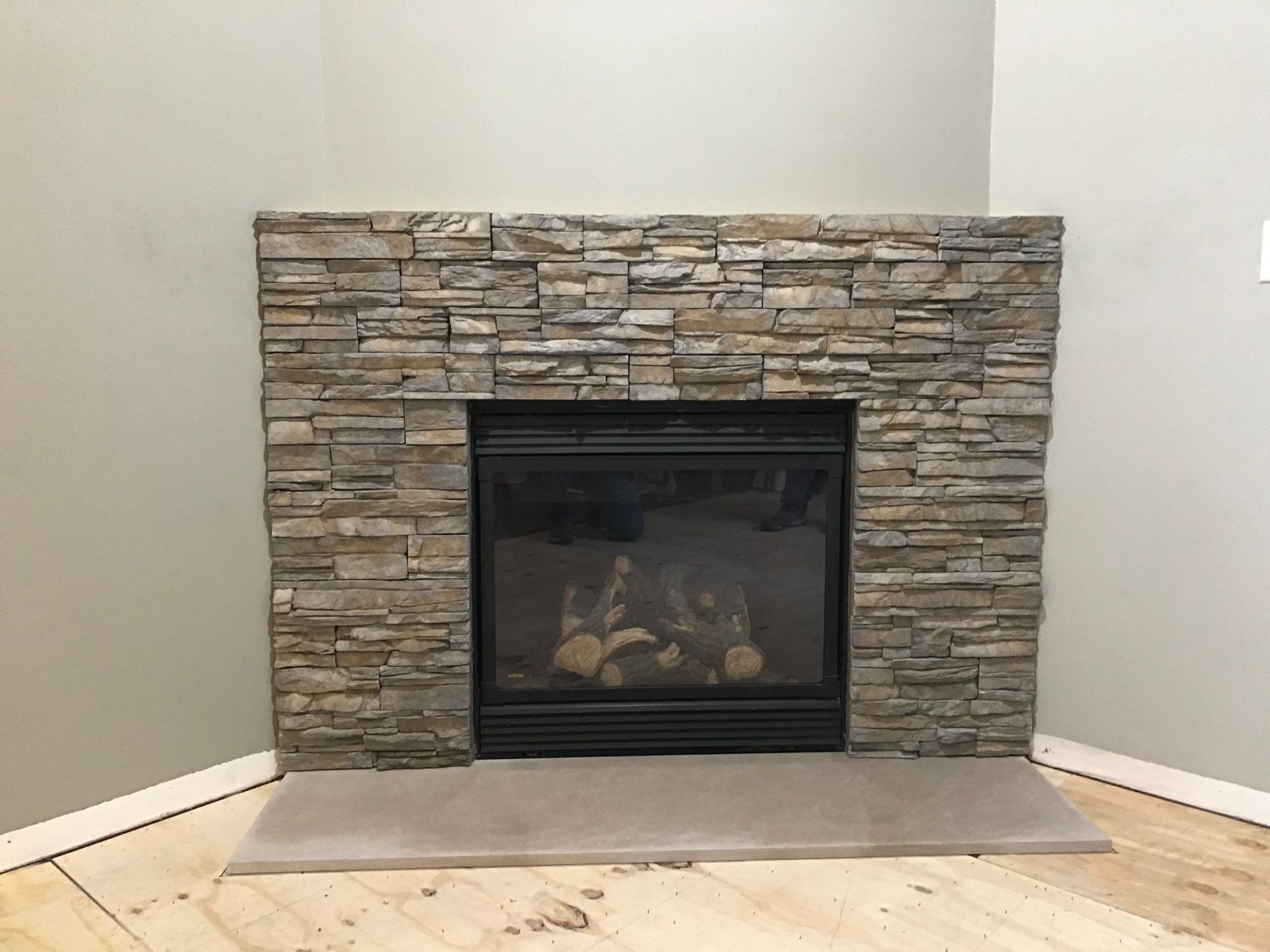 A corner fireplace with a dark glass firebox, stacked stone surround, and a flat concrete hearth on light wood flooring.