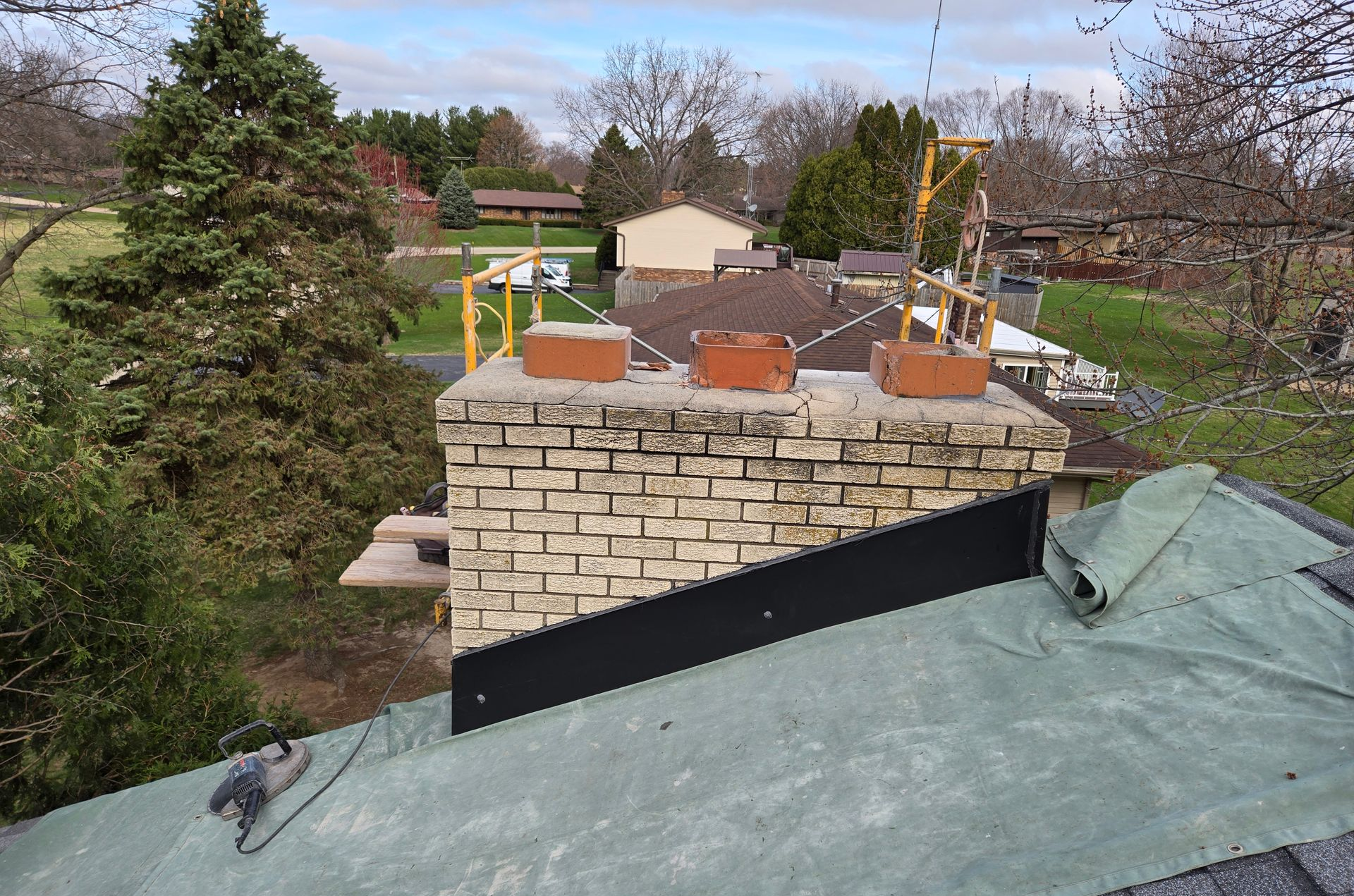 An elevated view of a brick chimney on a house roof under construction with green underlayment and black metal flashing.