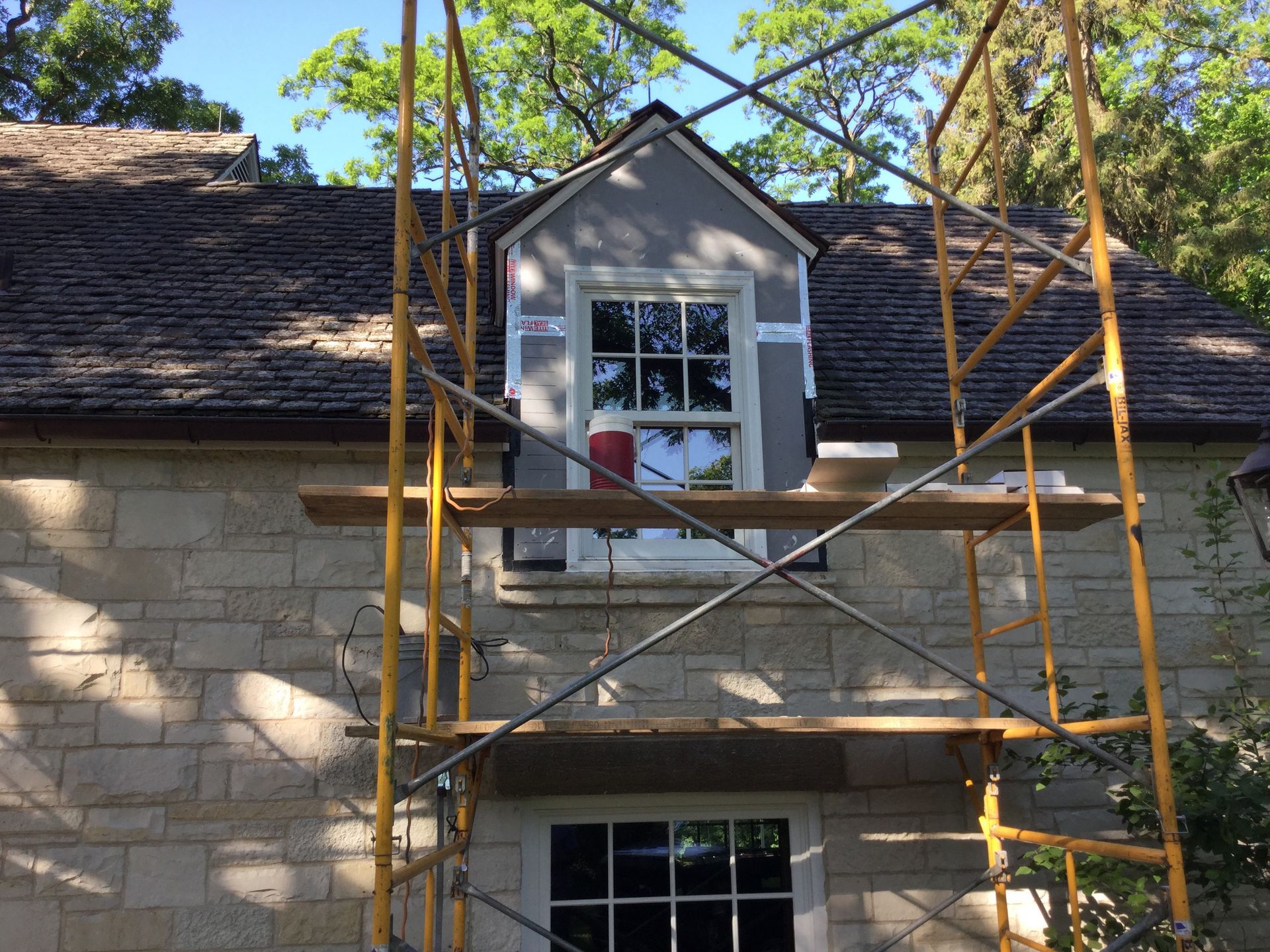 Yellow scaffolding stands in front of a stone house featuring a dormer window under repair, set against green trees.