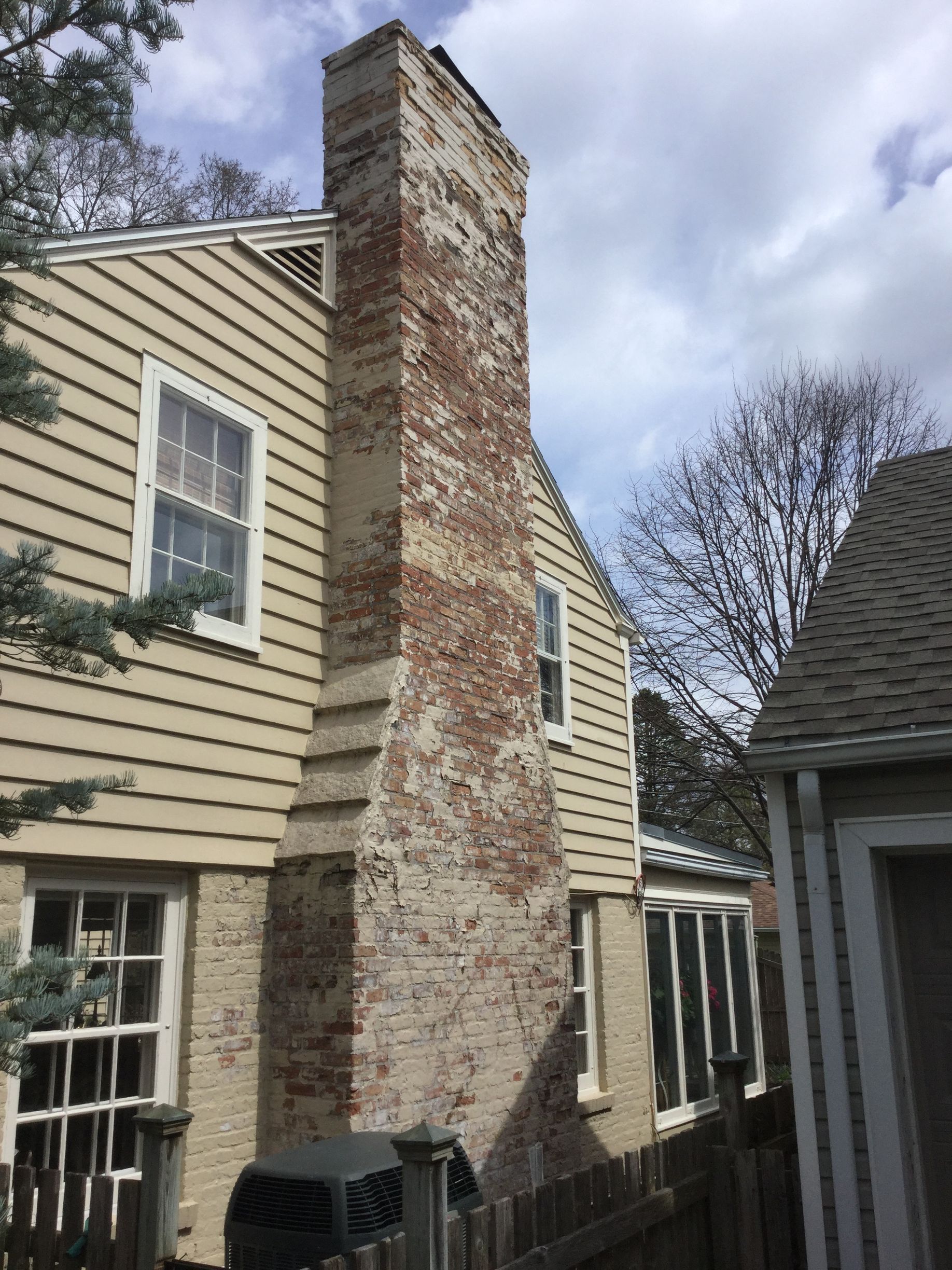 The side of a yellow-sided house featuring a tall, rustic brick chimney under a cloudy sky.