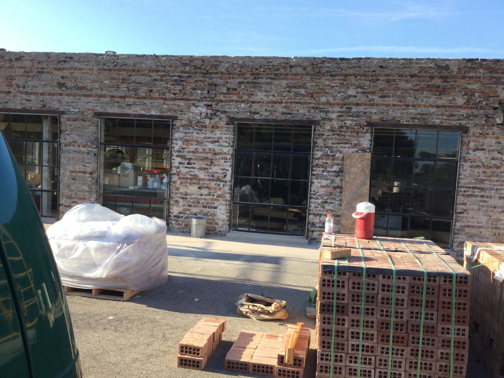 An outdoor construction site shows an old brick building with large glass windows, a pallet of bricks, and plastic wrap.