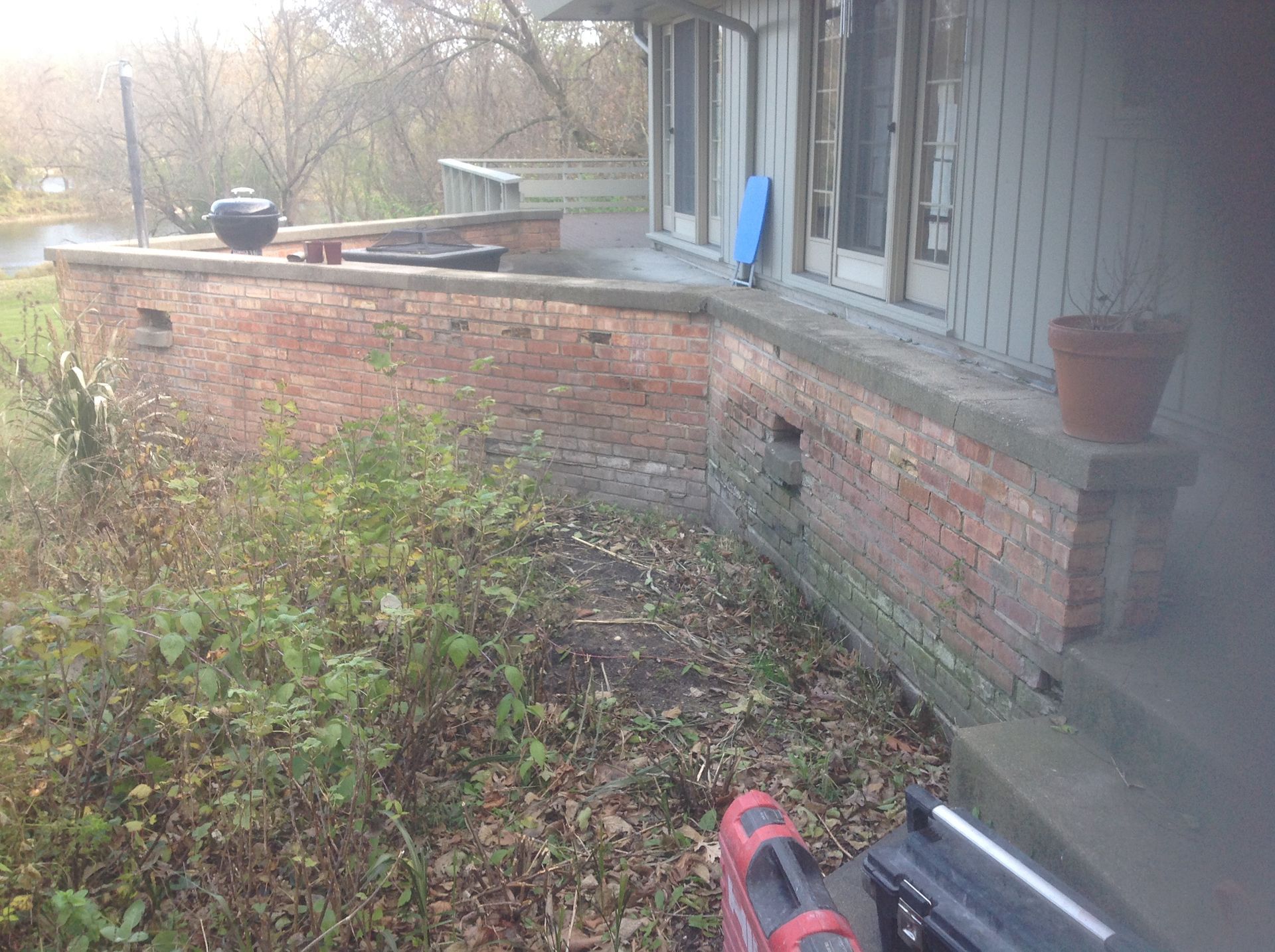 A brick patio or porch perimeter wall wraps around the side of a grey house, featuring a terracotta planter and foliage.
