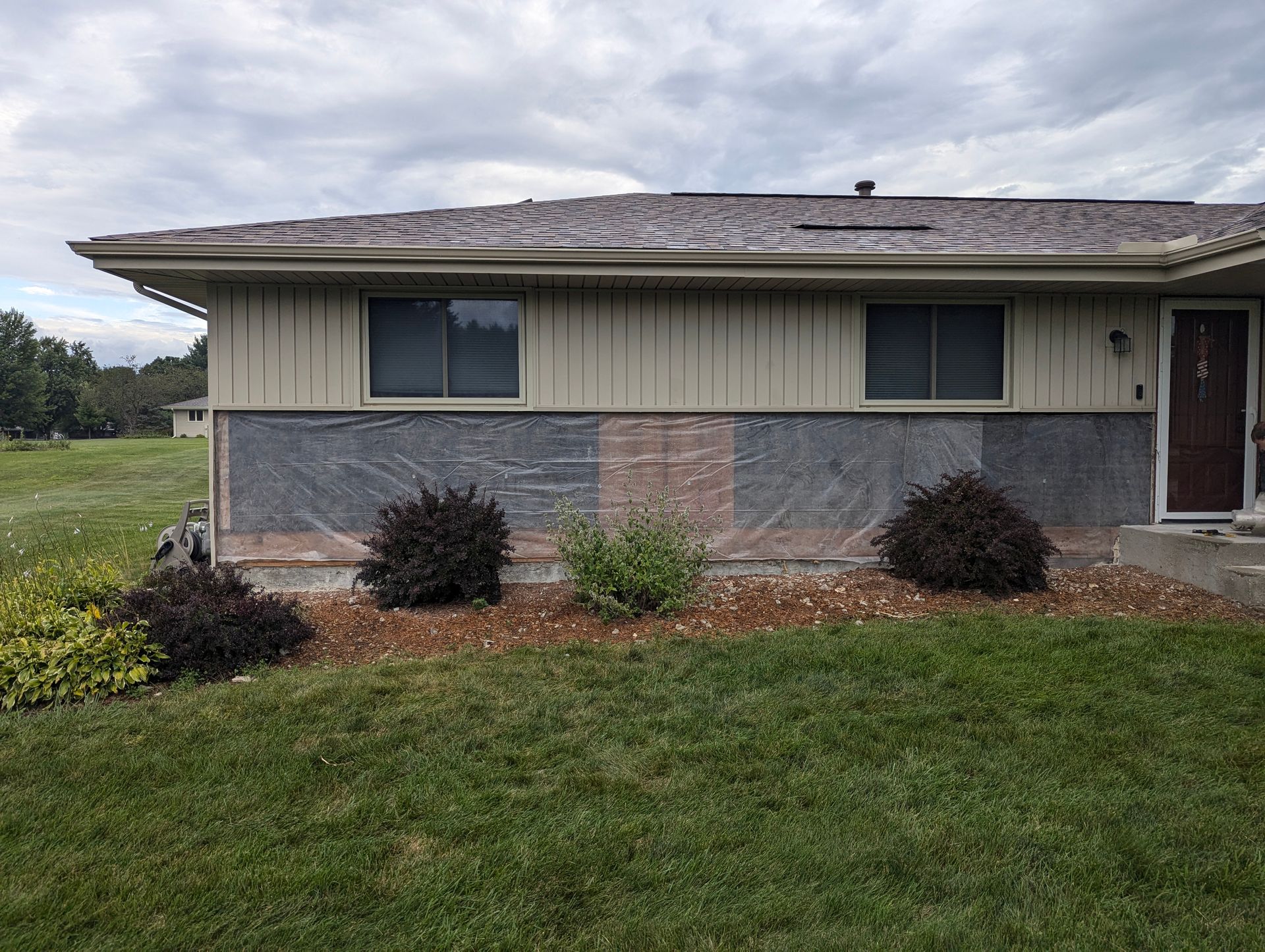 A single-story house with beige vertical siding over grey brick, damaged roofing, and landscaping in the front yard.