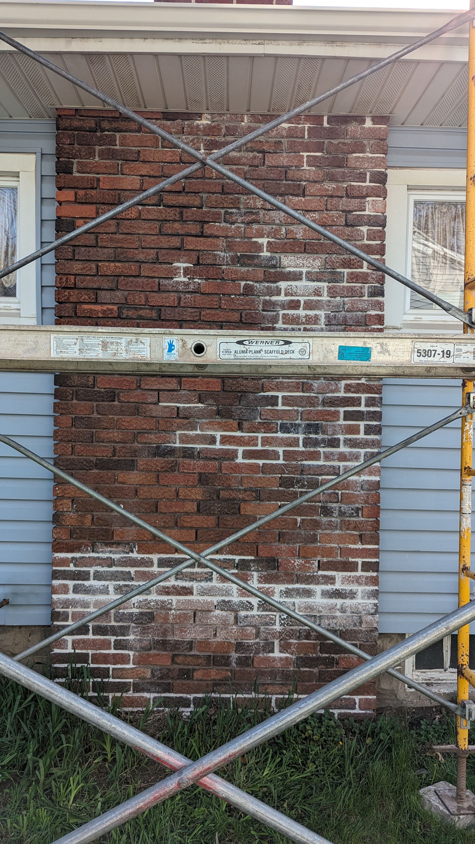 A brick fireplace chimney on the exterior of a house, partially obscured by yellow metal scaffolding.