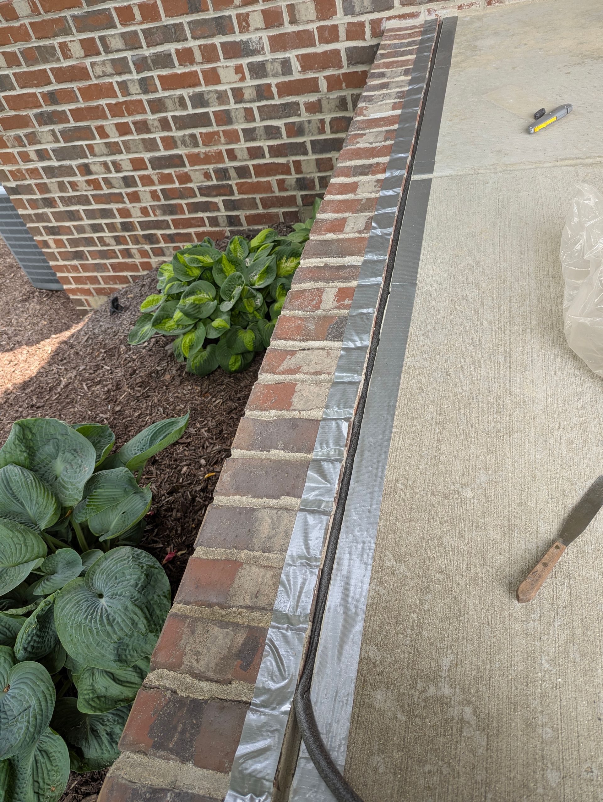 A brick garden border runs along a concrete patio, featuring a freshly applied gray sealant strip between the two surfaces.