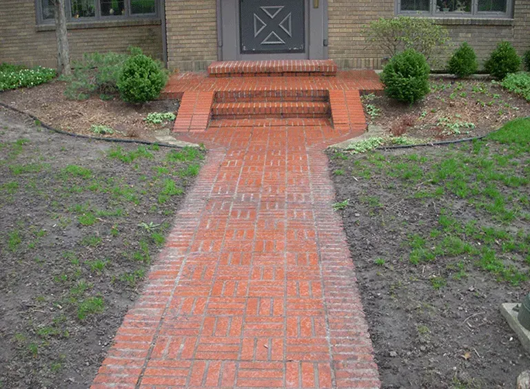 A red brick walkway leads to two brick steps at the entrance of a brick house with a dark, patterned front door.