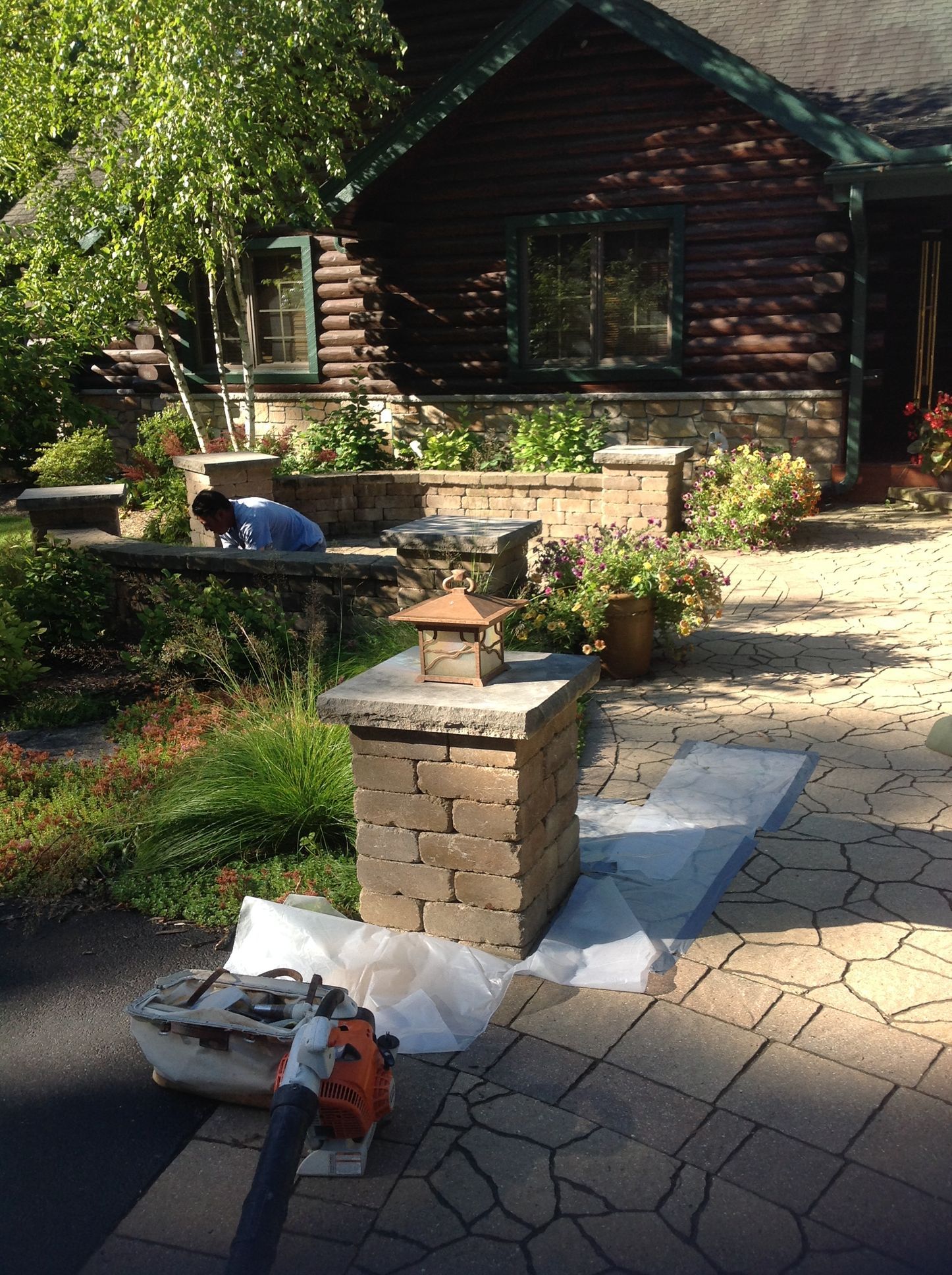 A person working on stone landscape pillars in front of a log cabin with a paved patio.
