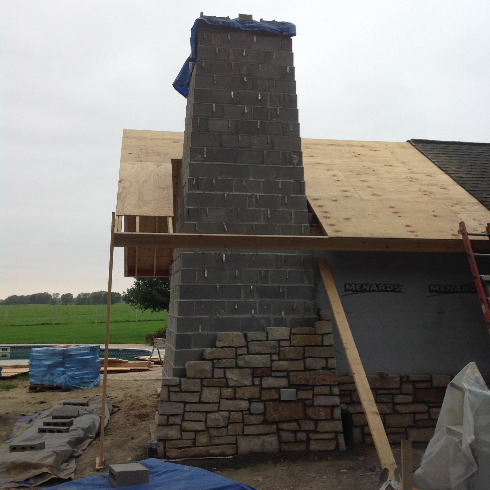 A gray concrete block chimney under construction, partially faced with natural stone, against a rural landscape.