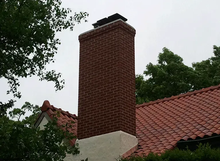 A brick chimney rises from a roof covered in red clay tiles, set against a backdrop of trees and a cloudy sky.