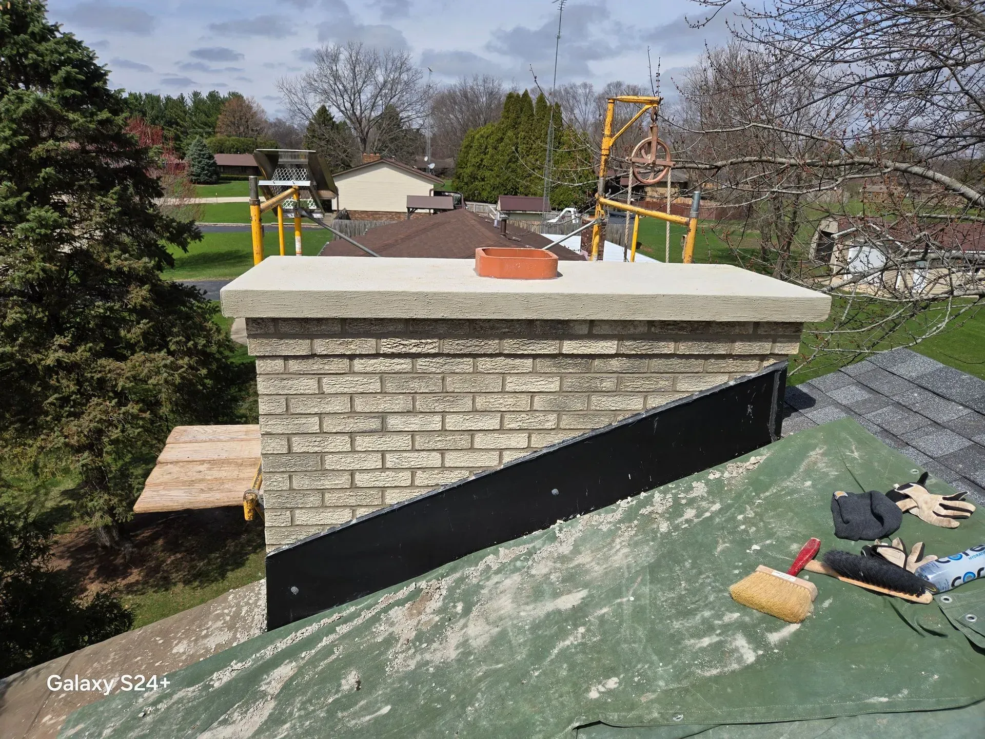 A brick chimney under construction on a roof, featuring a new concrete crown, metal flashing, and roofing materials nearby.