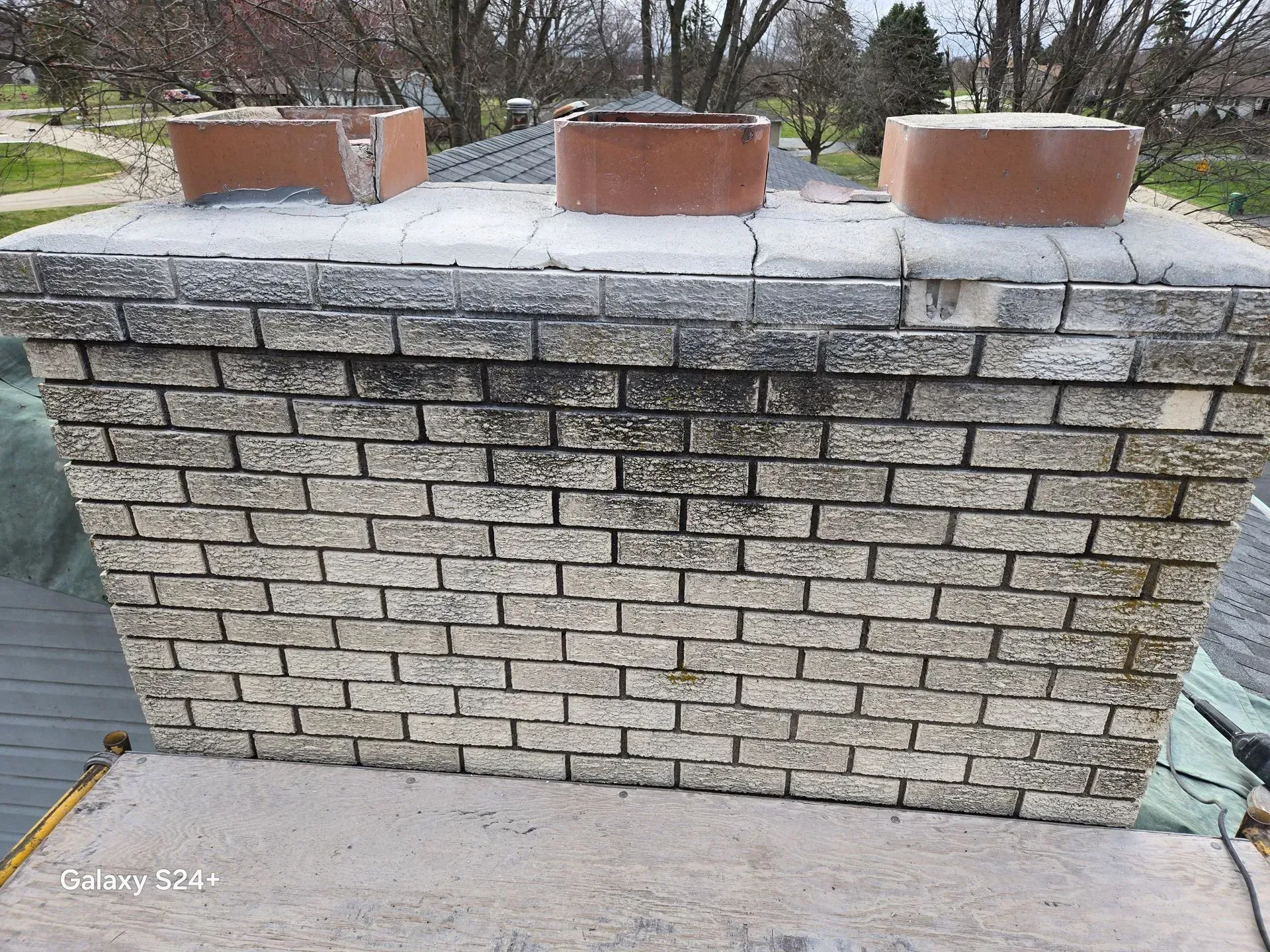 A rectangular masonry chimney with three terracotta flue liners on top, set against a blurred outdoor background.
