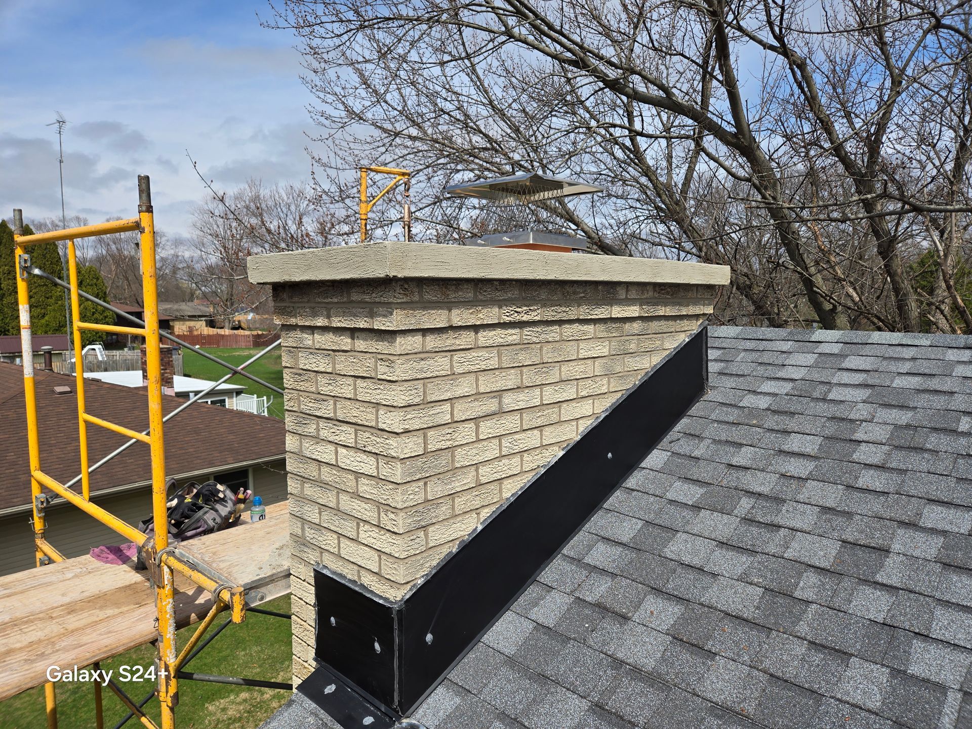 A tan brick chimney on a shingled roof with new black metal flashing installed, next to a yellow construction scaffold.
